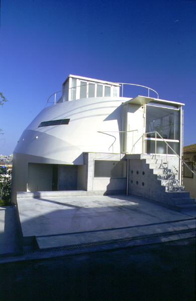 3 in 1 House このプロジェクトは、３住戸を共同化することによって、全体としておたがいのプライバシーを適切に確保しながら、戸建て感覚の環境的配慮がより可能な建築となっている。経済的メリットとしても、少々広い宅地の購入を可能にし、建設コストも抑えられるため、ほどほどに都心に近く、環境のよい既成住宅地の中で住宅を得ることができたものである。当然そういった敷地は、バブルがはじけて地価が下がったとはいえ、まだまだコスト面では負担が重すぎる。しかし一般的な宅地としては敬遠される傾斜地や変形地は、平坦地に比較すれば地価はかなり安く購入でき、しかも敷地の特性を生かした表現豊かな住まいの設計を可能にした。<br />この住宅のロケーションは、阪神間の少し山手にさしかかる既成住宅地で、前面道路から一層分落ち込んだ斜面地の敷地であり、各住戸の玄関へは２階部分からアプローチする。全体は80~90m²程度の３住戸を組み合わせ、外観上は３住戸の区分は一見わからない、ひとつのヴォリュームの塊の建築である。各住戸はプライベートな外部空間を確保するよう、１階はふたつの住戸を敷地の東西に２分させ、直接地面に接する庭をもつメゾネットタイプの住戸とし、最上階の住戸はペントハウスから屋上スペースを使用できるようにした。<br />またもっとも見晴らしのよい、視界の開けた南東部は、前面ガラスサッシュで開放させ、各住戸のパブリックスペースを配している。それに対し、北西側外壁の鉄板シェルターの建築形態は、この３住戸全体を殻のようにひとつに包み込み、住戸のプライバシーを確保し、また有機的なイメージと機械的な乗物のイメージをだぶらせ、ロマンを感じさせるような表現を試みようとしたものである。実際的にはこの鉄板シェルターの曲面は、北側隣地に影響する逆日影曲線によって、その形状は決定されている。<br />各住戸内部は、フレキシビリティを保つため、間仕切りを少なくし、全体を２分に大別し、パブリックとプライベートなスペースを対照的に対比させている。外部に開放的なパブリック空間と、内部を包み込むような閉ざされたプライベートな空間を、回遊的または連続するようにおたがいに結びつけている。<br />この周辺も阪神大震災の著しい被害を受けた住宅地域内である。この住宅は震災以前から計画は進めれ、震災時は工事中であったが、幸い被害はなかった。現在地域の復興は進んでいるが、高台になったこの敷地から見渡すかぎり、特色のないプレハブメーカーの住宅の建設ラッシュであり、そのスピードに驚くばかりである。しかし、より深刻な問題となっているのは、狭小宅地や４m未満の不適格道路に接した宅地で、住宅復興はいまだに遅れ、そのうえ住環境的な改善は見られない。この小規模共同化住宅の試みが、そのような住環境でのひとつの解決モデルにならないかと思っている。<br />（健康な住まいコンクール優秀賞　受賞）