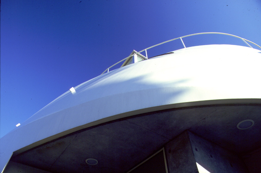 3 in 1 House このプロジェクトは、3住戸を共同化することによって、全体としておたがいのプライバシーを適切に確保しながら、戸建て感覚の環境的配慮がより可能な建築となっている。経済的メリットとしても、少々広い宅地の購入を可能にし、建設コストも抑えられるため、ほどほどに都心に近く、環境のよい既成住宅地の中で住宅を得ることができたものである。当然そういった敷地は、バブルがはじけて地価が下がったとはいえ、まだまだコスト面では負担が重すぎる。しかし一般的な宅地としては敬遠される傾斜地や変形地は、平坦地に比較すれば地価はかなり安く購入でき、しかも敷地の特性を生かした表現豊かな住まいの設計を可能にした。<br />この住宅のロケーションは、阪神間の少し山手にさしかかる既成住宅地で、前面道路から一層分落ち込んだ斜面地の敷地であり、各住戸の玄関へは2階部分からアプローチする。全体は80~90m²程度の3住戸を組み合わせ、外観上は3住戸の区分は一見わからない、ひとつのヴォリュームの塊の建築である。各住戸はプライベートな外部空間を確保するよう、1階はふたつの住戸を敷地の東西に2分させ、直接地面に接する庭をもつメゾネットタイプの住戸とし、最上階の住戸はペントハウスから屋上スペースを使用できるようにした。<br />またもっとも見晴らしのよい、視界の開けた南東部は、前面ガラスサッシュで開放させ、各住戸のパブリックスペースを配している。それに対し、北西側外壁の鉄板シェルターの建築形態は、この3住戸全体を殻のようにひとつに包み込み、住戸のプライバシーを確保し、また有機的なイメージと機械的な乗物のイメージをだぶらせ、ロマンを感じさせるような表現を試みようとしたものである。実際的にはこの鉄板シェルターの曲面は、北側隣地に影響する逆日影曲線によって、その形状は決定されている。<br />各住戸内部は、フレキシビリティを保つため、間仕切りを少なくし、全体を2分に大別し、パブリックとプライベートなスペースを対照的に対比させている。外部に開放的なパブリック空間と、内部を包み込むような閉ざされたプライベートな空間を、回遊的または連続するようにおたがいに結びつけている。<br />この周辺も阪神大震災の著しい被害を受けた住宅地域内である。この住宅は震災以前から計画は進めれ、震災時は工事中であったが、幸い被害はなかった。現在地域の復興は進んでいるが、高台になったこの敷地から見渡すかぎり、特色のないプレハブメーカーの住宅の建設ラッシュであり、そのスピードに驚くばかりである。しかし、より深刻な問題となっているのは、狭小宅地や4m未満の不適格道路に接した宅地で、住宅復興はいまだに遅れ、そのうえ住環境的な改善は見られない。この小規模共同化住宅の試みが、そのような住環境でのひとつの解決モデルにならないかと思っている。<br />(健康な住まいコンクール優秀賞 受賞)