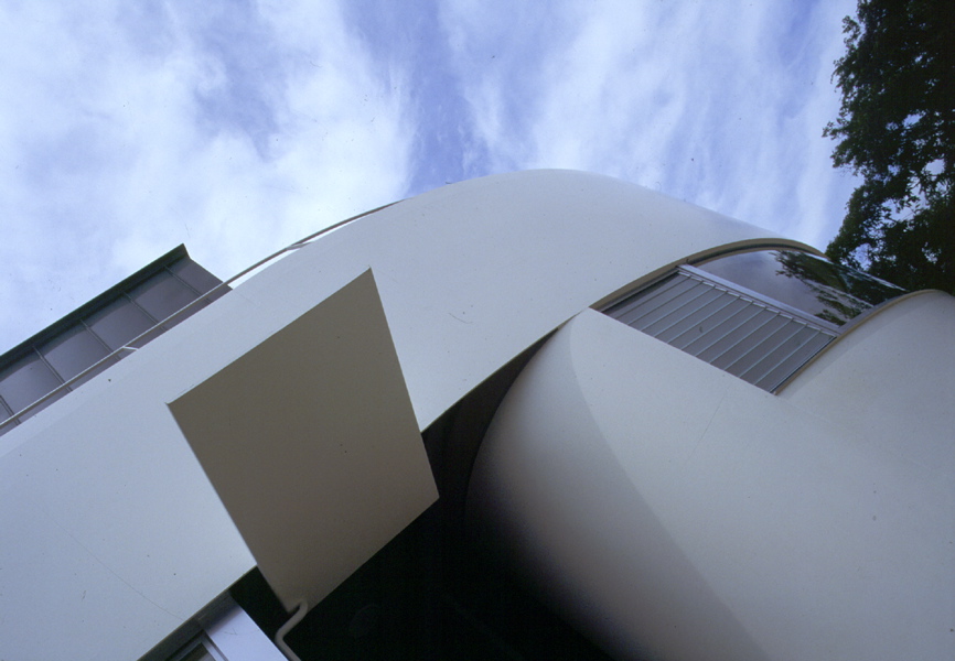 1/4 Circle House (東邸) この住宅の敷地は、六甲山系の東端・岩倉山の中腹にあり、周辺は東から南にかけて宅地開発をまぬがれた雑木林が山の斜面づたいに続いている。宅地ブロックの東南角というロケーションは、六甲山の優美な姿を中景として、遠景に遥か大阪湾をも見渡すことを可能にしている。このパノラミックな眺望が今回の住宅の性格を決定づけている。9mm厚、全長20mのスチールシートが、半径約 6mの曲率で敷地に沿ってほぼ90度曲げられ、この建物の外観を特徴づけている。この2枚の2次曲面は、構造からもち出すことで9.5mの無柱の横連窓を実現し、内部空間に開放的なパノラマ・ビスタを提供している。本来視角を遮へいするものである鉄板が図象的な解釈によって反転し、逆に開放的な開口部を印象づける役割を担っている。<br />一方、平面計画上においてもこの反転が意図されている。玄関からホールにかけて外部が内部に貫入し、中庭で再び垂直方向に外部へとつながる空間の連続は、入口から中庭まで床を同一材、壁を同色にすること、中庭立面をフラットに処理することで表れている。その内部空間によって囲われた外部(中庭)からは、内部(居間・キッチン・寝室1)を通してもうひとつの外部(パノラマ・ビスタ)を見通すことができる。同時に、機能コアとなるべき部分にヴォイドを配することで、周辺諸室が連結され、回遊する動線がつくり出される。また、中庭の透明なガラス・ボックスは、周辺環境の季節感をそこに投影し、住まいの内に陽光を導き入れる環境装置ともなっている。<br />そして建主の家族構成は2世帯なのだが、中庭を間に挟むことで、必ずしも明確な境界を設定することなく、緩やかな分離と共有を比較的簡単に実現している。寝室1と和室とを、居間と寝室2とを縦方向につなぐふたつのサーキュレーションは、2階のレベルで再び結合し上下層を回遊する動線となっている。さらに、中庭を中心として1階部分の西側を母親が、2階部分の北側を若夫婦が利用できるように計画され、中庭につながったオープンなキッチンと、パノラマ・ビスタをもつ居間というコモン・スペースを、共有することを可能にしている。<br />すなわち、プライバシーの確保と利便性、あるいは自然環境の導入と快適性という都市住宅における問題を、鉄板という材料と、中心に中庭を配置する方法で明快に解決しようとする試みである。<br /><br />