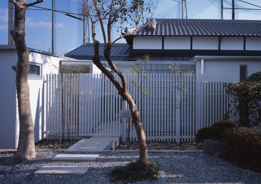 鉄の民家 この民家は、大阪近郊に残っていた貴重な民家である。これまで、幾度となく増改築が行われ、骨格と外観正面を除けば原型をとどめず、室内も新建材で改装されてしまっていた。残された資料も無く、建築主の記憶の聞き取りと建物調査から、増改築の痕跡を探り原型の復元に努めた。建設年代は、屋根裏から御弊が発見され、上棟は大正14年(1925年)11月とわかり、そして、古い土壁に貼られた昭和4年の新聞片も発見された。その壁面はガラスで覆い「痕跡」として保存した。全体は、改修と補強を行ったこの原型となる民家部分と、新たに改修築したスチール・シートで包まれた寝室とサニタリー部分、そして、和室の庭に設けられた小さな離家の茶室の3つに区分することができる。そして、この民家の木造部分とスチール・シートによって改修築された部分の間に、トップライトのある通り土間廊下を土間のリビング+キッチンを設けた。このインターフェイスとしての土間空間を挿入することによって、環境調整を伴う光と風抜きのパッシブ的役割も担い、また、空間の新旧のメリハリと分節しながらも全体の各室を結び付け、昔乍らの和の生活と現代生活をうまく融合し、蘇生できたと考えている。<br /><br />スチール・シートによって改修築された部分は、t=9mmのスチール・シートで寝室とサニタリーを包むように囲っている。長筒状のフラットルーフの形状で、木造の民家部分とは対照的に車両が停車したように見える。既存の柱梁を取り替え、スチール・シートによって補強し、既存の柱間と合わせた半間間隔(960mm)にアングル型鋼をリブ状に建て、それを溝型鋼で門型につなぎ連続させている。この工法は、鉄船舶の居住部などを建造する工法と同じで、建築が柱梁に頼らない、より軽快なリブ構造によってモノコック建築の試みである。そして、土間廊下は、構造的緩衝体となり、民家部分の木造とこのモノコックの異種構造を力の伝達の少ないトップライトで繋いでいる。全体のディテールは、空間に突起部分のないできるだけ平滑でスッキリと格納した、何でない納まりとなるように考えた。