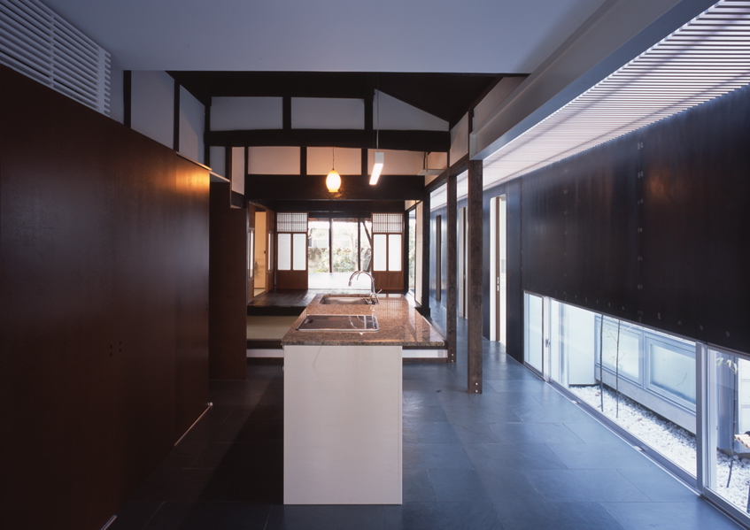 鉄の民家 この民家は、大阪近郊に残っていた貴重な民家である。これまで、幾度となく増改築が行われ、骨格と外観正面を除けば原型をとどめず、室内も新建材で改装されてしまっていた。残された資料も無く、建築主の記憶の聞き取りと建物調査から、増改築の痕跡を探り原型の復元に努めた。建設年代は、屋根裏から御弊が発見され、上棟は大正14年(1925年)11月とわかり、そして、古い土壁に貼られた昭和4年の新聞片も発見された。その壁面はガラスで覆い「痕跡」として保存した。全体は、改修と補強を行ったこの原型となる民家部分と、新たに改修築したスチール・シートで包まれた寝室とサニタリー部分、そして、和室の庭に設けられた小さな離家の茶室の3つに区分することができる。そして、この民家の木造部分とスチール・シートによって改修築された部分の間に、トップライトのある通り土間廊下を土間のリビング+キッチンを設けた。このインターフェイスとしての土間空間を挿入することによって、環境調整を伴う光と風抜きのパッシブ的役割も担い、また、空間の新旧のメリハリと分節しながらも全体の各室を結び付け、昔乍らの和の生活と現代生活をうまく融合し、蘇生できたと考えている。<br /><br />スチール・シートによって改修築された部分は、t=9mmのスチール・シートで寝室とサニタリーを包むように囲っている。長筒状のフラットルーフの形状で、木造の民家部分とは対照的に車両が停車したように見える。既存の柱梁を取り替え、スチール・シートによって補強し、既存の柱間と合わせた半間間隔(960mm)にアングル型鋼をリブ状に建て、それを溝型鋼で門型につなぎ連続させている。この工法は、鉄船舶の居住部などを建造する工法と同じで、建築が柱梁に頼らない、より軽快なリブ構造によってモノコック建築の試みである。そして、土間廊下は、構造的緩衝体となり、民家部分の木造とこのモノコックの異種構造を力の伝達の少ないトップライトで繋いでいる。全体のディテールは、空間に突起部分のないできるだけ平滑でスッキリと格納した、何でない納まりとなるように考えた。