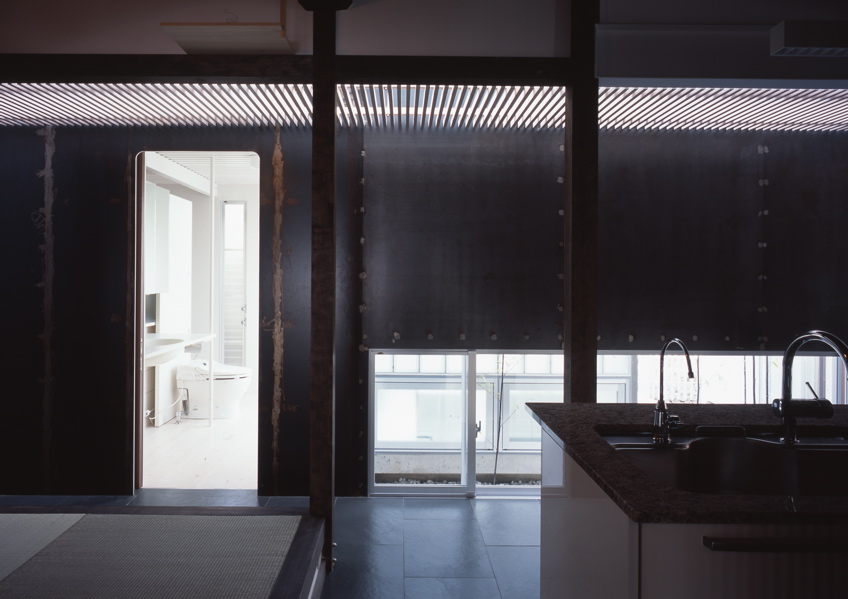 鉄の民家 この民家は、大阪近郊に残っていた貴重な民家である。これまで、幾度となく増改築が行われ、骨格と外観正面を除けば原型をとどめず、室内も新建材で改装されてしまっていた。残された資料も無く、建築主の記憶の聞き取りと建物調査から、増改築の痕跡を探り原型の復元に努めた。建設年代は、屋根裏から御弊が発見され、上棟は大正14年(1925年)11月とわかり、そして、古い土壁に貼られた昭和4年の新聞片も発見された。その壁面はガラスで覆い「痕跡」として保存した。全体は、改修と補強を行ったこの原型となる民家部分と、新たに改修築したスチール・シートで包まれた寝室とサニタリー部分、そして、和室の庭に設けられた小さな離家の茶室の3つに区分することができる。そして、この民家の木造部分とスチール・シートによって改修築された部分の間に、トップライトのある通り土間廊下を土間のリビング+キッチンを設けた。このインターフェイスとしての土間空間を挿入することによって、環境調整を伴う光と風抜きのパッシブ的役割も担い、また、空間の新旧のメリハリと分節しながらも全体の各室を結び付け、昔乍らの和の生活と現代生活をうまく融合し、蘇生できたと考えている。<br /><br />スチール・シートによって改修築された部分は、t=9mmのスチール・シートで寝室とサニタリーを包むように囲っている。長筒状のフラットルーフの形状で、木造の民家部分とは対照的に車両が停車したように見える。既存の柱梁を取り替え、スチール・シートによって補強し、既存の柱間と合わせた半間間隔(960mm)にアングル型鋼をリブ状に建て、それを溝型鋼で門型につなぎ連続させている。この工法は、鉄船舶の居住部などを建造する工法と同じで、建築が柱梁に頼らない、より軽快なリブ構造によってモノコック建築の試みである。そして、土間廊下は、構造的緩衝体となり、民家部分の木造とこのモノコックの異種構造を力の伝達の少ないトップライトで繋いでいる。全体のディテールは、空間に突起部分のないできるだけ平滑でスッキリと格納した、何でない納まりとなるように考えた。