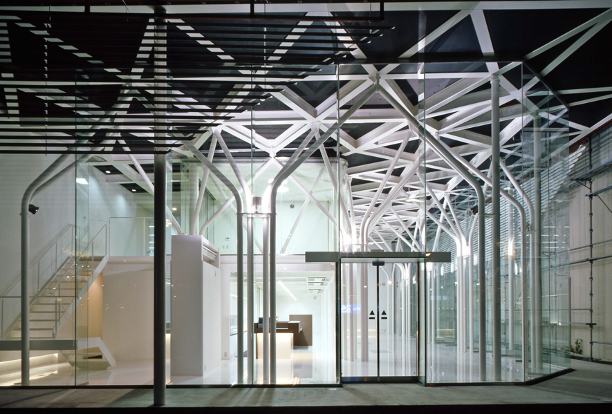 ドコモショップ彦根店・長浜店・能登川店 一般的には、店舗型であり、このDOCOMO Shopは、郊外1棟型の比較的大きな携帯専用ショップで、希なケースである。全体は、オープンスペースの待ち合いとカウンタースペース、バックオフィス、そして、2階に広めの会議室と小会議室がある。ショップ部分は、地域のパブリックスペースとして捕らえ、市民(利用者)の快い居場所として提供出来ればと考えた。公共性は地域と繋がる方法である。そして、このショップ部分の2層の大空間からなるヴォリュームは、単調なボイド空間に成らぬように、魅力ある空間の提供がテーマのであった。抽象的な人工の森をイメージし開放的な空間を想定し、ダイナミックでエレガントな構造体と意匠性が一体と成った空間体感のあるスペースを求めた。 (木村博昭)<br /><br /> 新しいイメージを放っているDOCOMO Shopの特徴は,細い柱が林立する開放的な空間 である.この空間は待合スペースのため,壁やブレースが配置できず,可能な限り細い部材であることが要求された.そこで,地震力は全て事務スペース部のラーメンで 負担し,待合スペースは鉛直荷重と積雪荷重に耐えられるように設計することで,φ 114.3パイプの林が実現した.待合スペース上部の屋根の地震力を事務スペース部に伝 えるため,屋根のトラスは剛接にした.また,丸パイプを曲げて加工した家形フレー ムは単体では非常に弱く,わずかな鉛直荷重しか支えられない.そこで,家形フレー ムが集まる位置に厚いダイヤフラムを配置し,家形フレームを強化した.このダイヤ フラムは,頂部から鉛直荷重を受けた家形フレームが広がるのを拘束する効果もある.また,非常に細い部材で座屈が心配だが,立体骨組解析を行い立体的な座屈の検 討も行った. <br /> (京都工芸繊維大学 森迫/金尾構造研究室 金尾 伊織)<br />