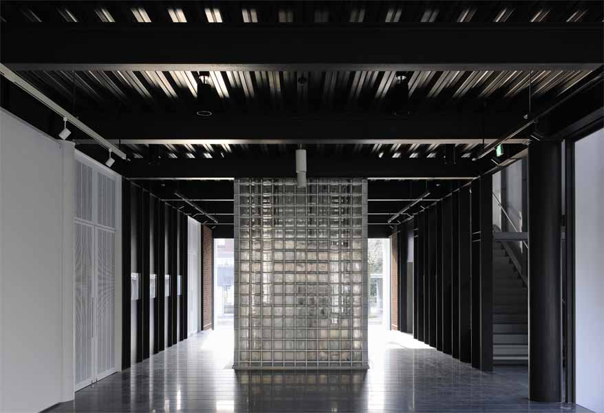 京都工芸繊維大学創立60周年記念館 60周年記念館は、シンボル的存在として大学中央口前に建つ。大学キャンバス計画に基づき、既存校舎の軸線に合わせ、既存樹木帯を残した外部道路との間に位置している。全体は、一階にメモリアルホール、ギャラリーと記念講義室があり、二階に大小のセミナーホールとそのホワイエラウンジがある。そして、一部構内の外構塀を取払い、直接外部道路からの使用可能な学外に開いた施設である。<br />記念館の構想にあたり、時間をテーマに記憶と時を静止させた時代を再確認する静的な建築であると捕らえた。建築は、過去から継承され失われる技術もあり、当然ながら機能では定まらず、感性、記憶、象徴等と多重的要素で成立している。<br />本大学創設当時の本野清吾設計による校舎は、やや重量感のあるRC造の建築であるが、しかし、W. Gropiusのドイツ工作連盟の透明感のあるFagus FactoryやBauhaus Buildingを想起させる新時代の建築であり、一時代の大学のシンボルとしてあり、当時のデザインスクールとして新たな近代デザインへの可能性を具現且つ体感的に示したものであった。<br />現代建築は、存在性が薄れつつある。しかしながら、建設に携わった職人達の無名の熟練した手の痕跡や建築素材により、建築そのものに命を与え、その空間体感が五感を呼び起こし、人に感動と敬意を促す。此れまで出会ったすばらしい建築は、それらが身体にダイレクトに伝わるものであった。そして、その時代の文化の結晶として残るのである。ここでは、そんな建築への思いと取り組み、空間の素材とディテールを熟考し、現代性の表現として鉄を使い、創設期の本野清吾のRC造の校舎と対称させた。<br />また、その表現として、空間の初原的な型としてのボルト架構の、2つの異なる相対によって、エレガント性とダイナミズム、そして、空間の格式と品格、更には、未来への可能性を暗示出来ればと考えた。